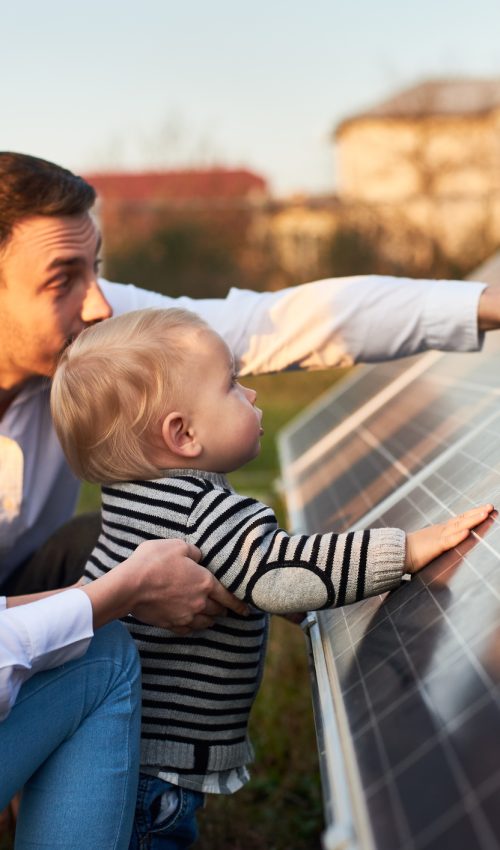 Side close-up shot of a young modern family with a little son getting acquainted with solar panel on a sunny day, green alternative energy concept